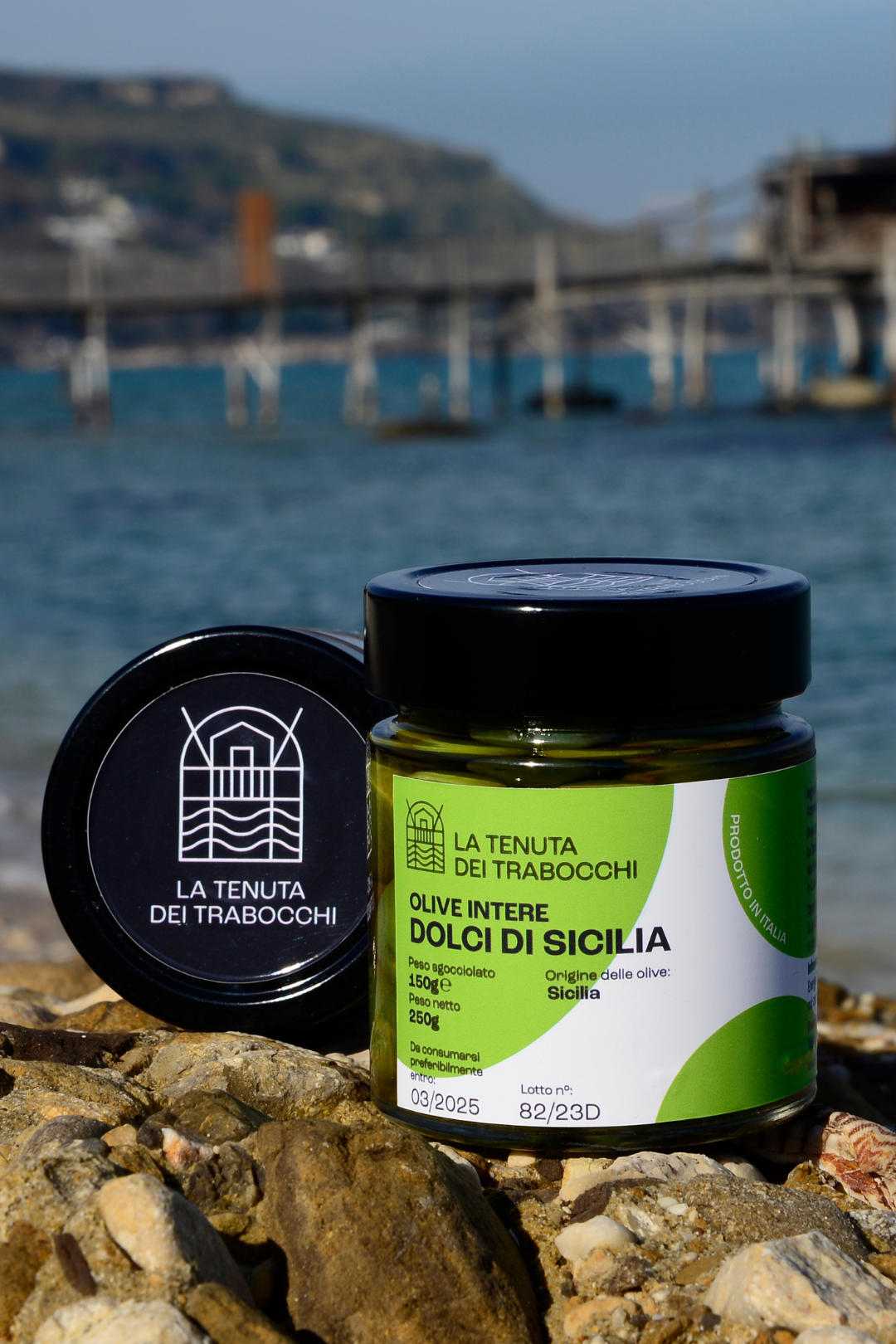 Vaso in vetro contenente olive intere e dolci di Sicilia della Tenuta Dei Trabocchi adagiato su una roccia con mare in fondo della costa Dei Trabocchi ed un trabocco