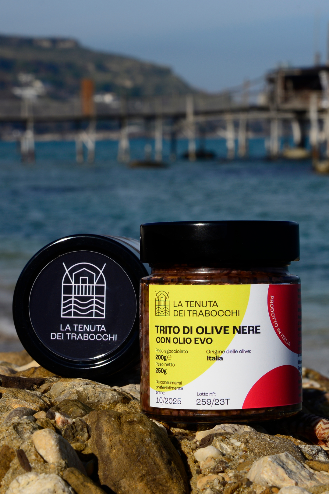 Vaso in vetro contenente trito di olive nere con olio extravergine della Tenuta Dei Trabocchi adagiato su una roccia con mare in fondo della costa Dei Trabocchi ed un trabocco