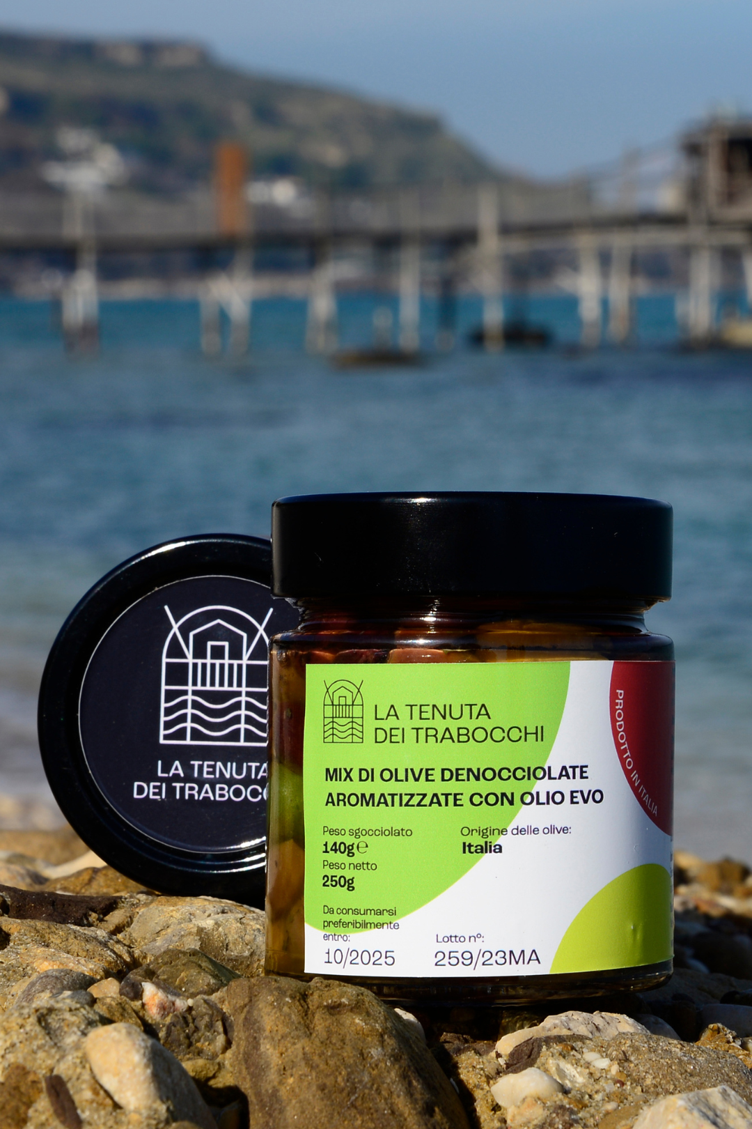 Vaso in vetro contenente mix di olive denocciolate aromatizzate della Tenuta Dei Trabocchi adagiato su una roccia con mare in fondo della costa Dei Trabocchi ed un trabocco
